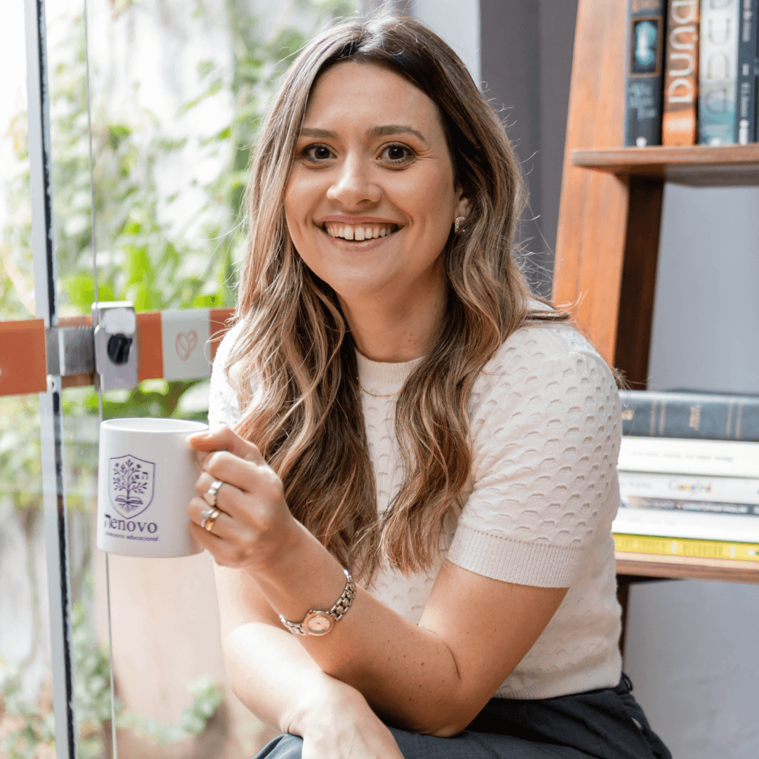Carol Darós sorrindo segurando uma caneca, em frente a uma estante de livros